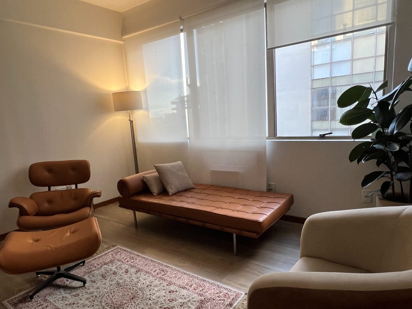 Sala de atendimento do consultório, com divã e poltrona de couro caramelo, luminária de chão acesa, tapete persa e janelas com persianas claras