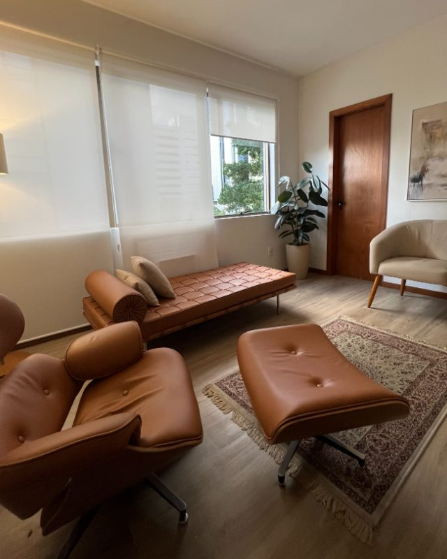 Vista ampla da sala, com divã, pufe de couro, poltrona bege e quadro abstrato na parede