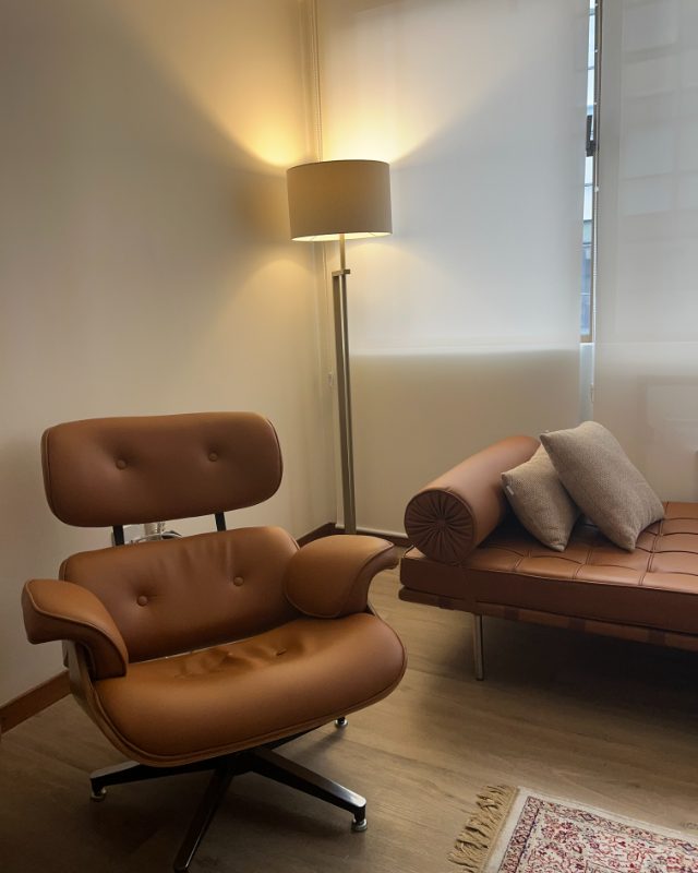 Poltrona de couro caramelo em primeiro plano, com a luminária acesa e o divã ao fundo